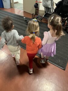 Three children are holding hands while walking through a hall.